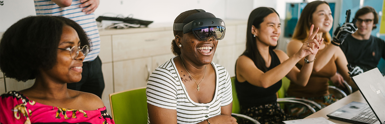 Lachende Studentin mit VR-Brille in einer Reihe von anderen Studierenden an einem Tisch